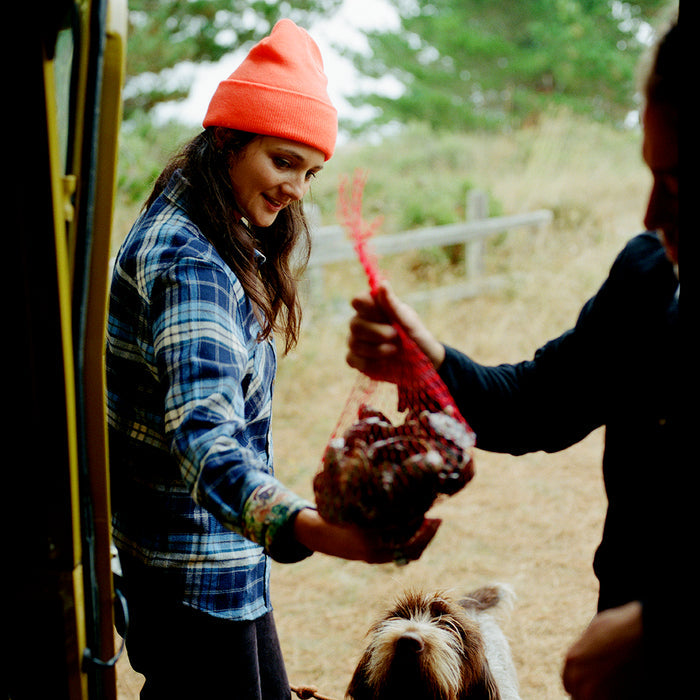 A woman in an orange beanie smiles, wearing Pladra’s Women's Every Day Elite Flannel Shirt in Bighorn Blue by a fence.