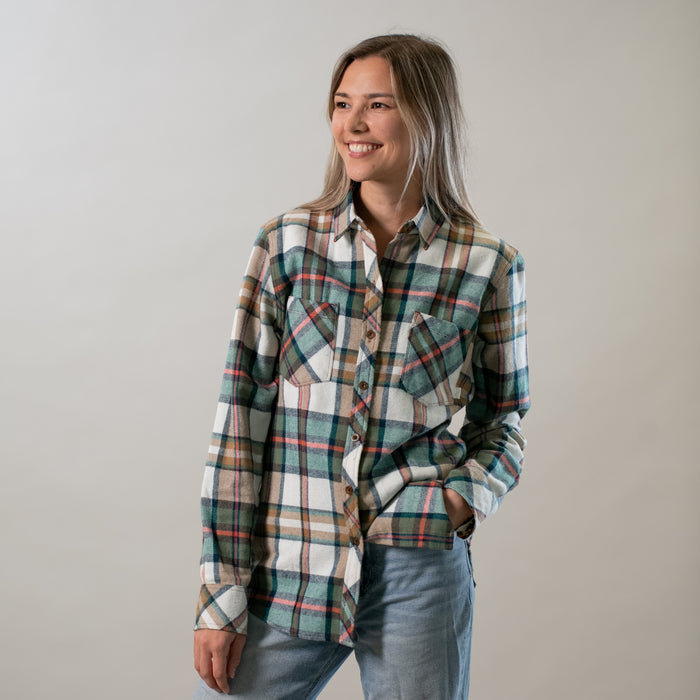 Smiling woman wears Pladra Women's Every Day Elite Flannel Shirt in Coyote Tan and jeans, hand in pocket, plain background.