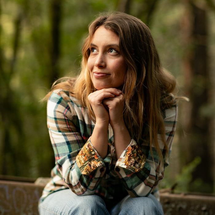 A woman in a Pladra Women's Every Day Elite Flannel Shirt in Coyote Tan smiles outdoors with a forest background.