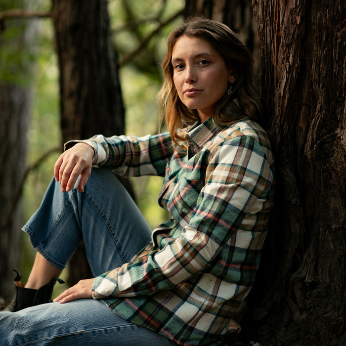 A woman in a Pladra Women's Every Day Elite Flannel Shirt–Coyote Tan sits calmly against a tree, facing the camera.