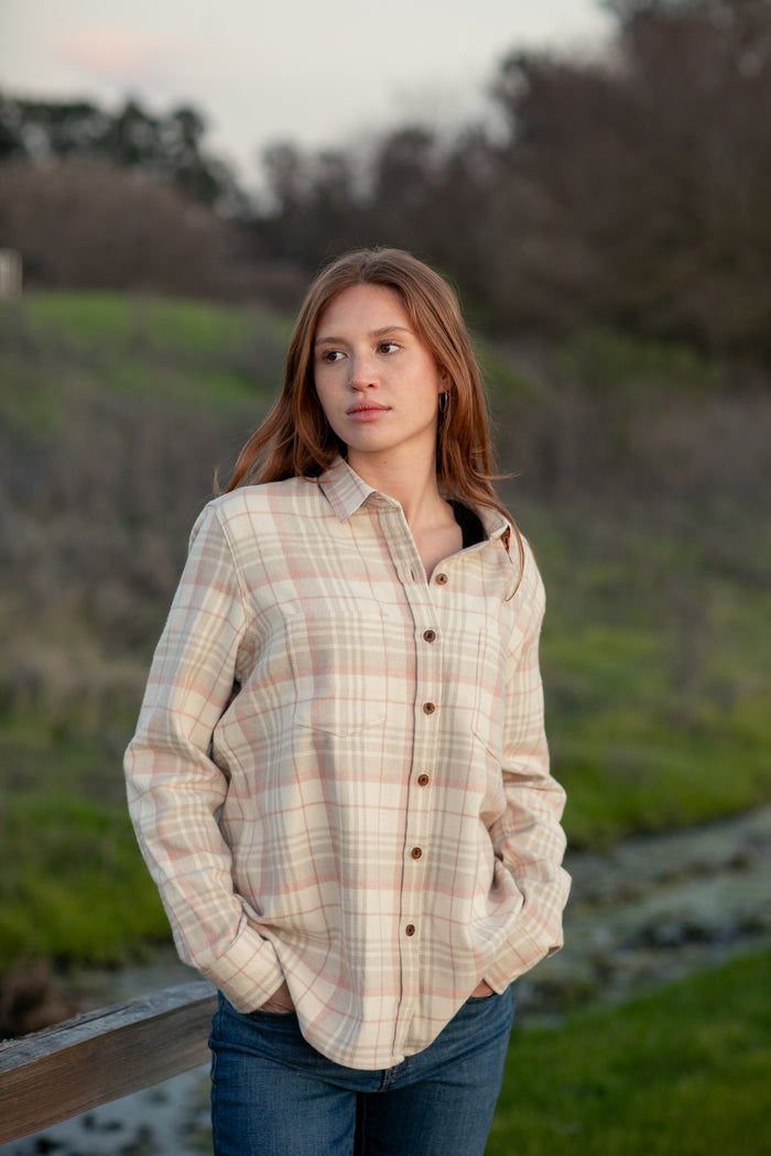A young woman with long brown hair, wearing the Women's Every Day Elite Flannel Shirt in Lighthouse White and blue jeans, stands outdoors on grassy ground with trees and a cloudy sky, hands in her pockets and looking to the side.