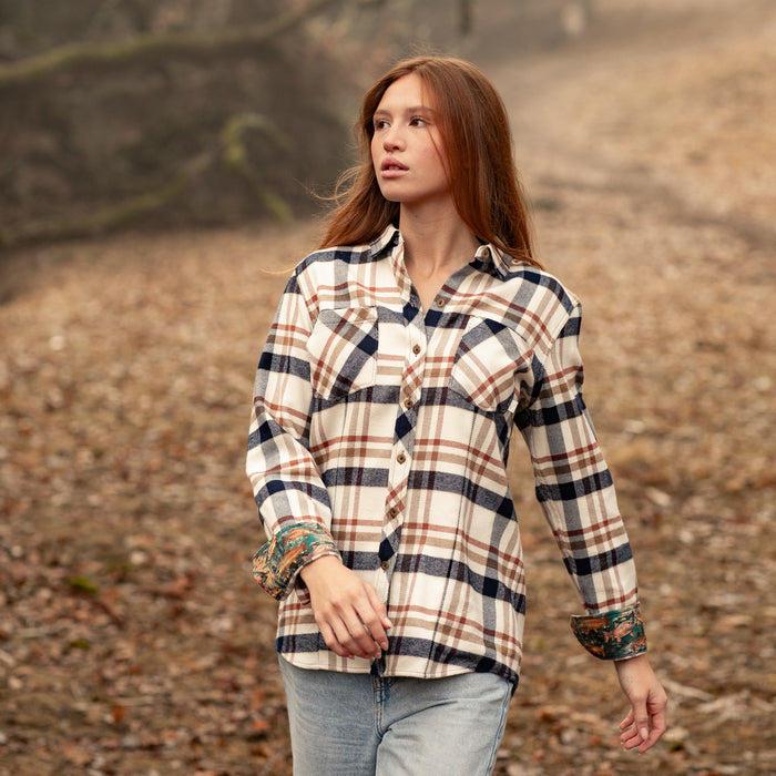 A woman with long reddish hair, wearing the Women's Every Day Elite Flannel Shirt in Whitefish Tan, walks outdoors on a leaf-strewn path, surrounded by blurred trees. She gazes to the side with a thoughtful look.