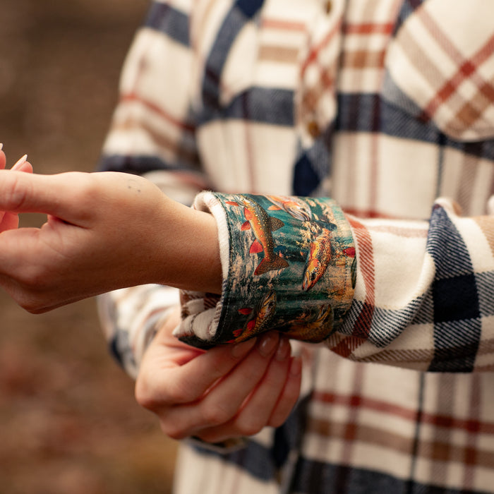 A woman adjusts the cuff of the Women's Every Day Elite Flannel Shirt-Whitefish Tan, revealing a colorful fish-patterned inner fabric. The blurred background highlights its premium cotton quality and unique details.