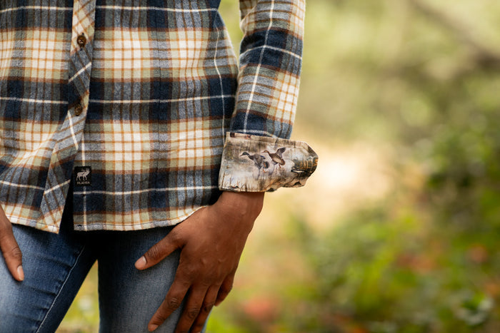 A woman wears the Women's Every Day Flannel Shirt in Banff Blue, a premium cotton shirt with a hunting dog print and rolled-up sleeves, paired with blue jeans and standing outdoors against lush greenery.