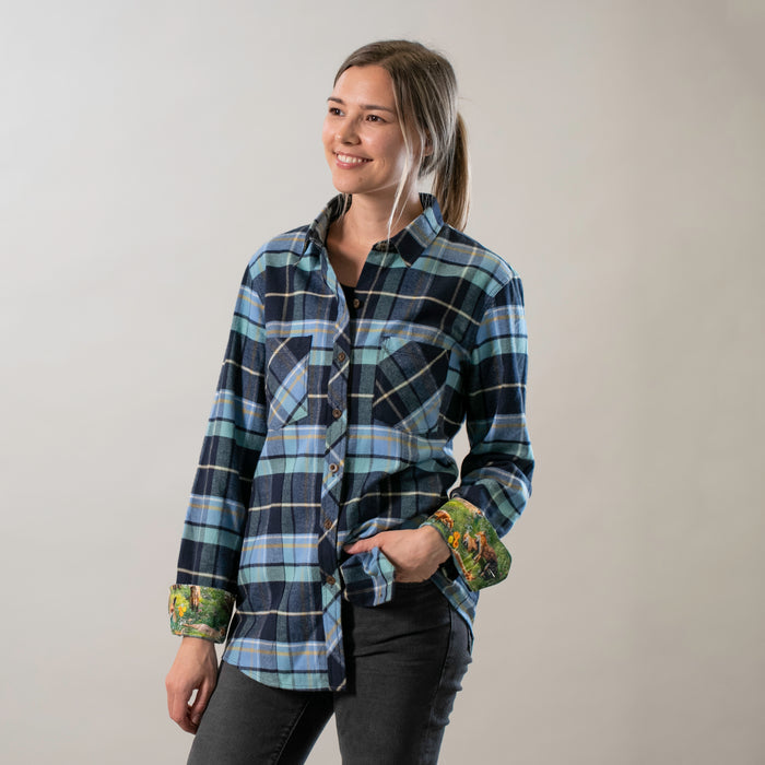 A woman with long hair tied back is smiling against a plain background, wearing the Women's Every Day Flannel Shirt- Bay Blue with rolled-up sleeves and dark pants.