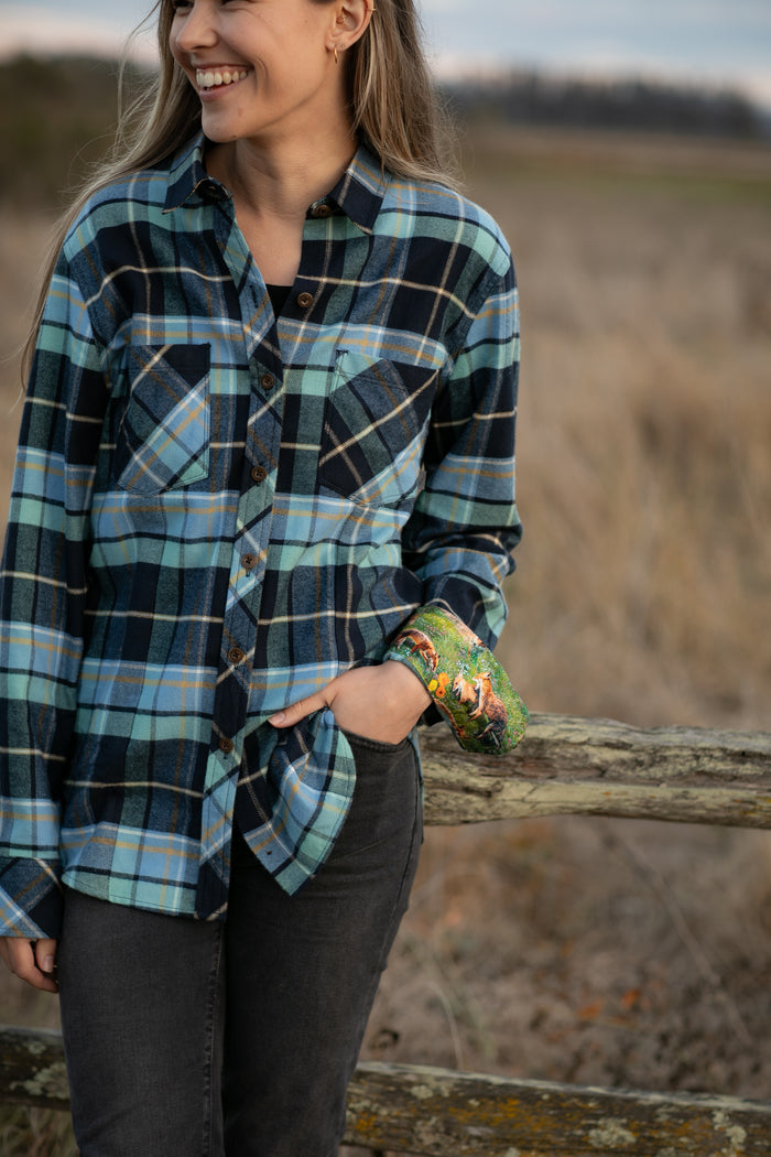 A woman stands outdoors by a wooden fence, smiling, in the Women's Every Day Flannel Shirt - Bay Blue and black jeans. Her left hand with a colorful cast is in her pocket, with a grassy field in the background.