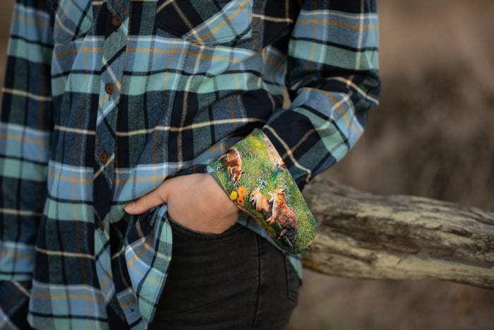 A person wears the Women's Every Day Flannel Shirt in Bay Blue with the cuff turned up, revealing a colorful fox design, as they stand outdoors with a hand in their pocket, leaning on a wooden rail.