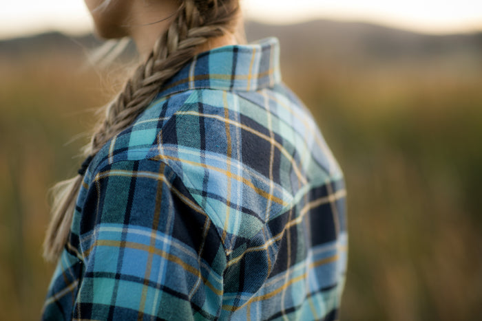 A close-up shows someone with a loose braid wearing the Women's Every Day Flannel Shirt in Bay Blue, standing outdoors with grassy fields and hills blurred in the background. The person's face is not shown.
