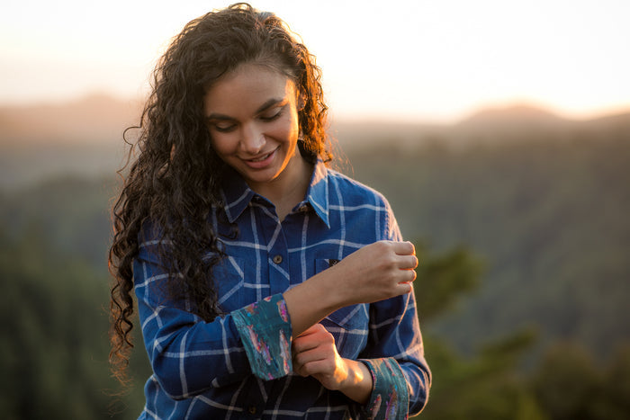 Smiling woman in Pladra's Women's Every Day Flannel Shirt—Billings Blue rolls up her sleeve outdoors.