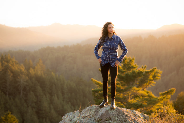 A woman in a Pladra Women's Every Day Flannel Shirt - Billings Blue stands on a rock overlooking a sunset valley.