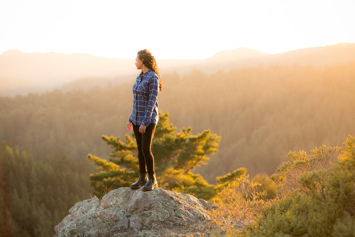 Woman in a Pladra Women's Every Day Flannel Shirt in Billings Blue stands on a rock, overlooking a sunset-lit forest.