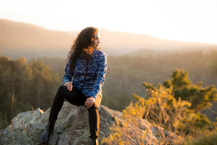 A woman in a Pladra Women's Every Day Flannel Shirt—Billings Blue sits on a rock, gazing at a sunset forest and mountains.