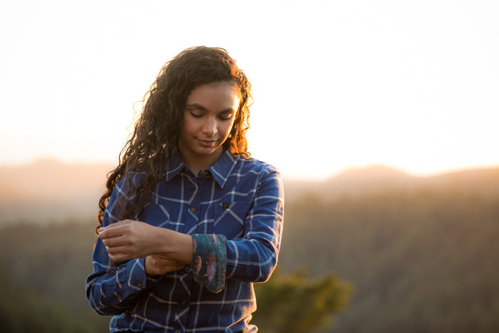 Woman in Pladra Women's Every Day Flannel Shirt–Billings Blue rolls up her sleeve outdoors at sunset.