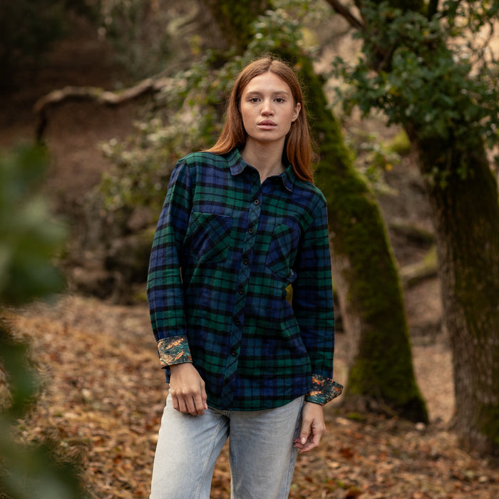 A woman wears the Pladra Women's Every Day Flannel Shirt in Bolinas Blue and jeans, standing among trees and leaves.