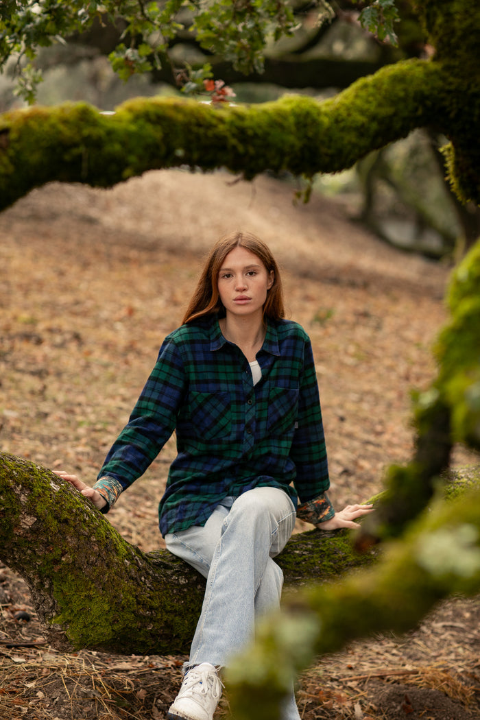 A woman in a Pladra Women's Every Day Flannel Shirt—Bolinas Blue sits on a mossy branch in the woods, facing the camera.