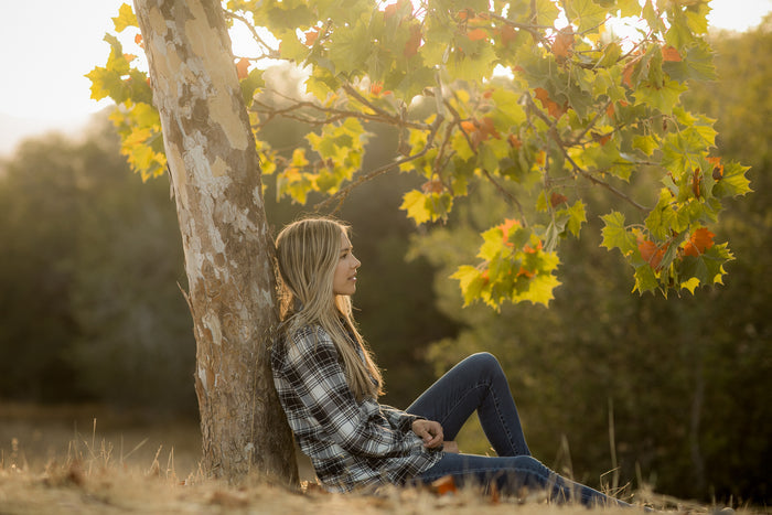 Young woman in a Pladra Women's Every Day Flannel Shirt-Butte Blue sits by a tree amidst sunlit autumn leaves.