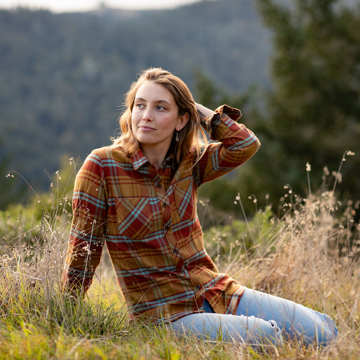 A woman in the Women's Every Day Flannel Shirt—Cottonwood Orange and jeans sits in a grassy field, gazing to the side with her hand in her hair. Blurred forested hills can be seen in the background.