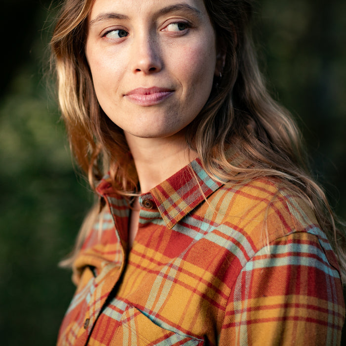 A woman with long, wavy hair wears the Women's Every Day Flannel Shirt in Cottonwood Orange plaid, made from premium cotton, and gazes off to the side, smiling softly outdoors in natural light.