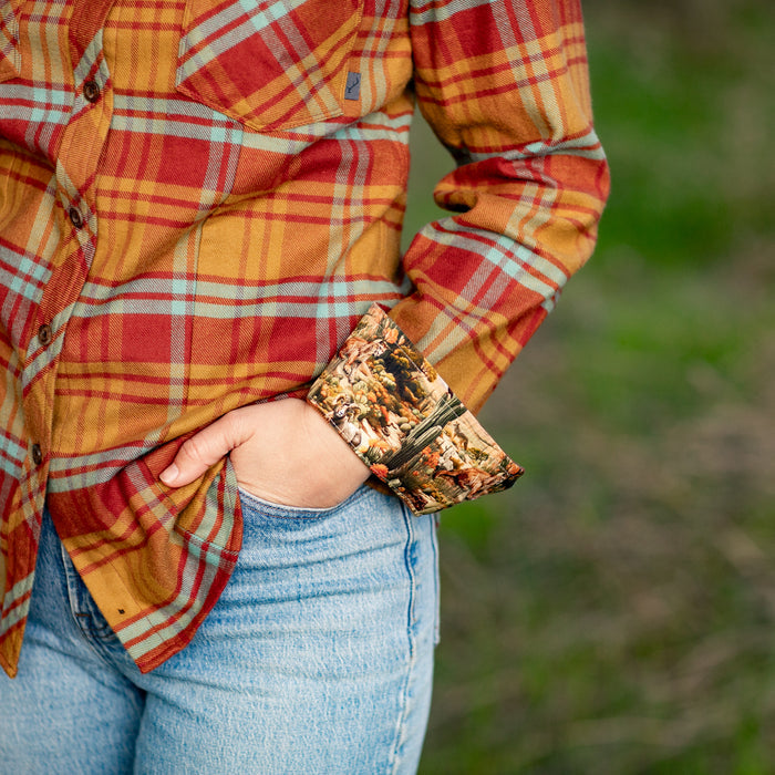 A woman outdoors stands in green grass, one hand in her pocket, wearing blue jeans and the Women's Every Day Flannel Shirt–Cottonwood Orange: a red and yellow plaid cotton shirt with rolled sleeves revealing a printed lining.