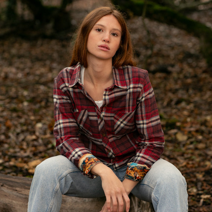 A woman in a Pladra Women's Every Day Flannel Shirt - Harvest Red and jeans sits on a log in the forest, looking calm.