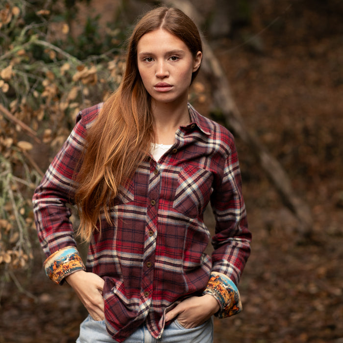 A woman with long red hair wears the Pladra Women's Every Day Flannel Shirt in Harvest Red, standing outdoors among trees.