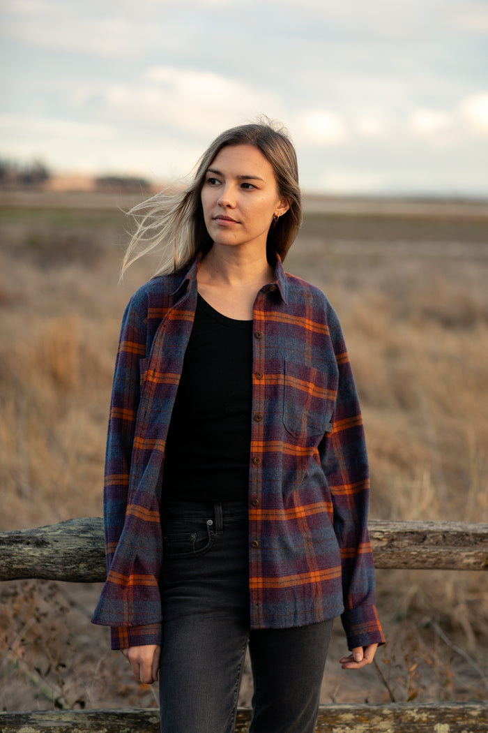 A woman in a Pladra Women's Every Day Flannel Shirt- Portsmouth Blue Heather stands outdoors by a wooden fence.