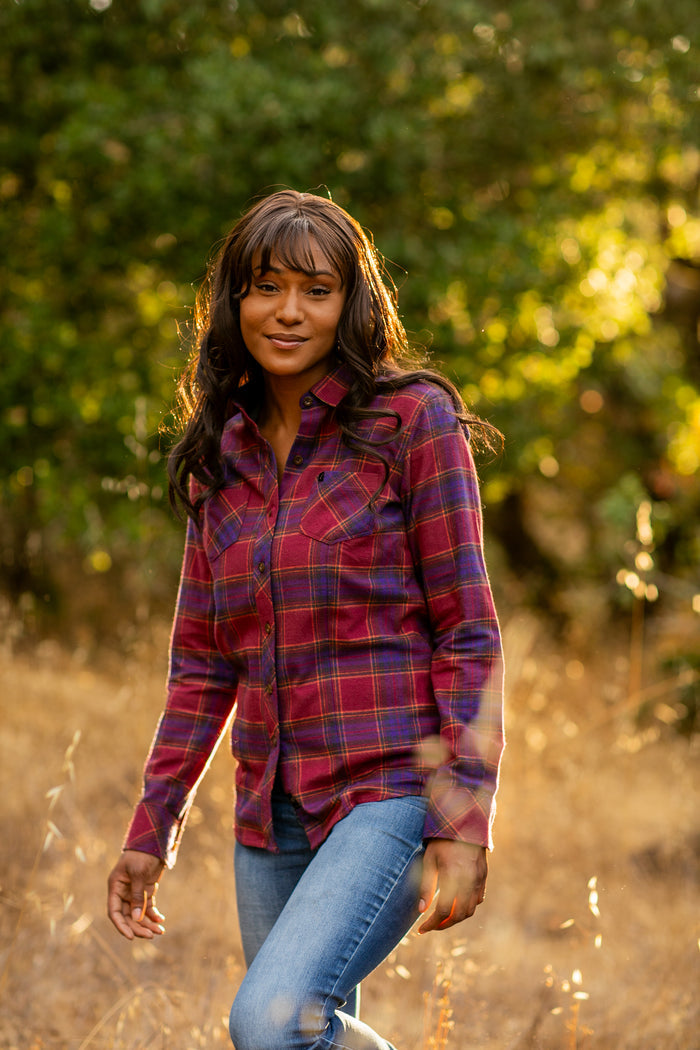 A woman in a Pladra Women's Every Day Flannel Shirt—RK Plum Red and jeans stands outside in a grassy, sunlit field.