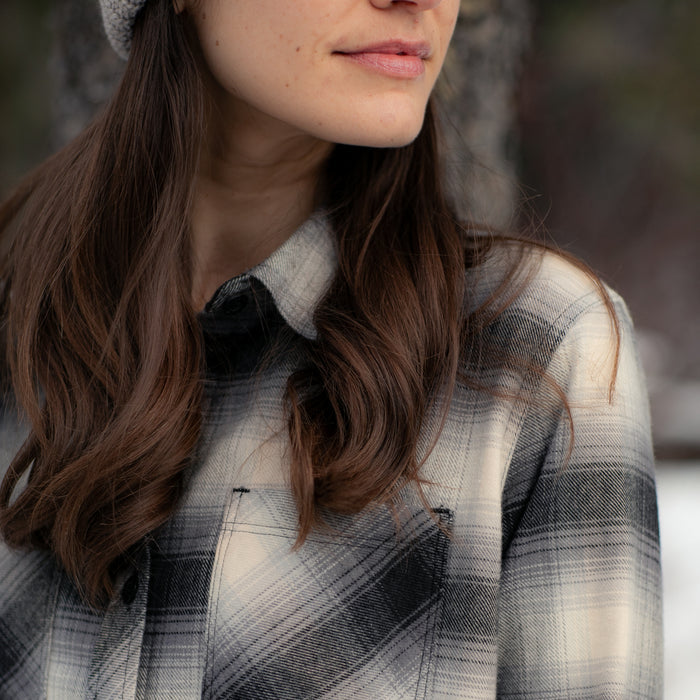 A woman with long brown hair enjoys the outdoors in a forest, wearing the Women's Every Day Flannel Shirt—Sterling Grey Heather. Only her lower face and upper torso in the cotton flannel are visible.