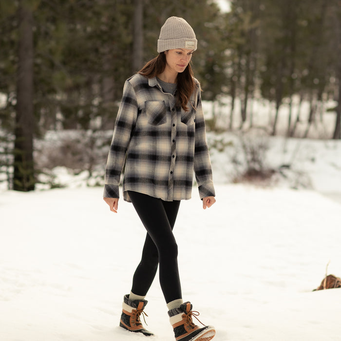A woman wearing the Women's Every Day Flannel Shirt in Sterling Grey Heather, black leggings, winter boots, and a knit beanie walks outdoors on snowy ground with pine trees in the background.