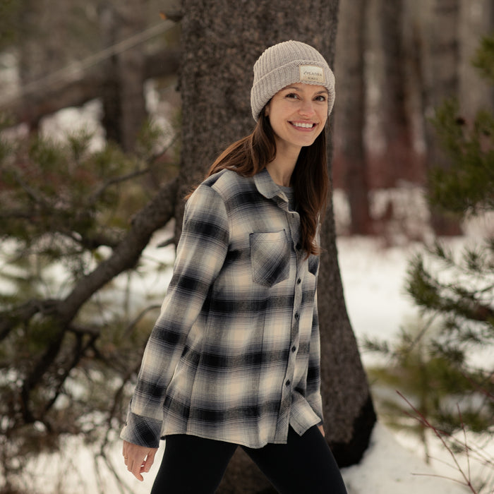 A woman in a beige knit beanie and the Women's Every Day Flannel Shirt—Sterling Grey Heather smiles while walking outdoors in a snowy forest, with pine trees and a tree trunk visible in the background.
