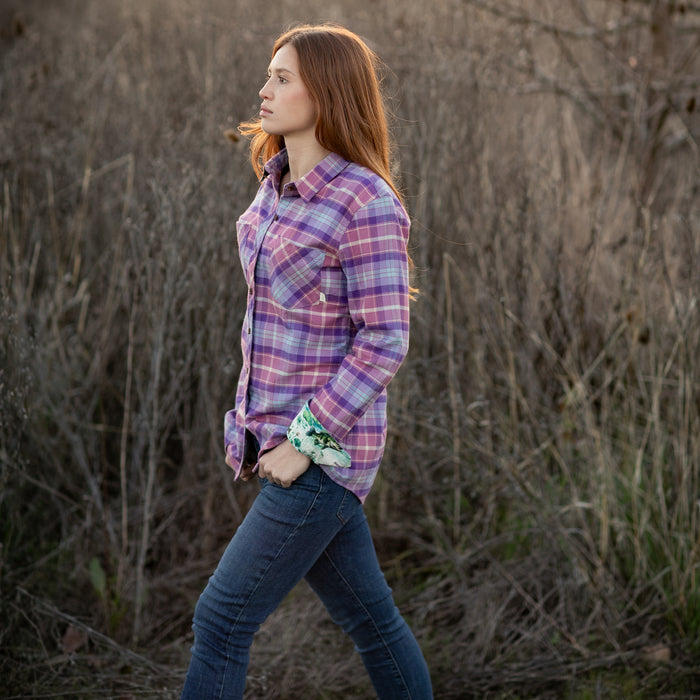 A woman with long red hair wears Pladra's Women's Every Day Flannel Shirt in Wildflower Pink and jeans outdoors.