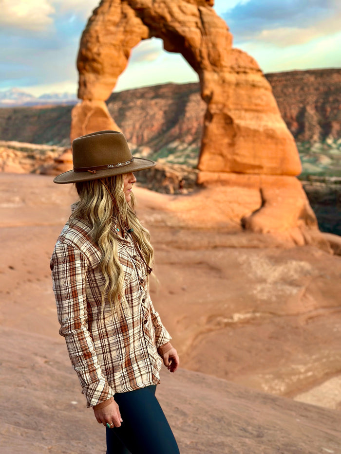 A woman wearing the Women's Fireside Flannel in Cattail Tan stands on rocky terrain with Utah's Delicate Arch in the background, under warm sunlight.