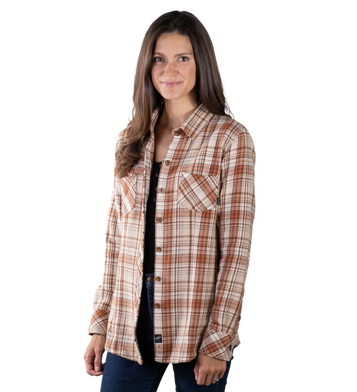 A woman with long brown hair smiles at the camera, wearing the Women's Fireside Flannel in Cattail Tan over a black top and dark jeans, standing against a white background.