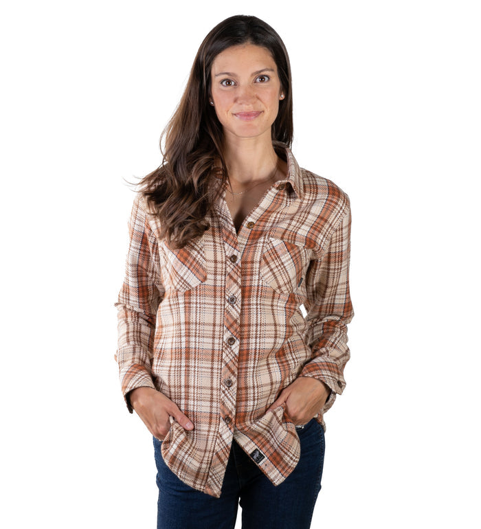 A woman with long brown hair wears the Women's Fireside Flannel in Cattail Tan and dark jeans, standing facing forward with her hands in her pockets and smiling against a white background.