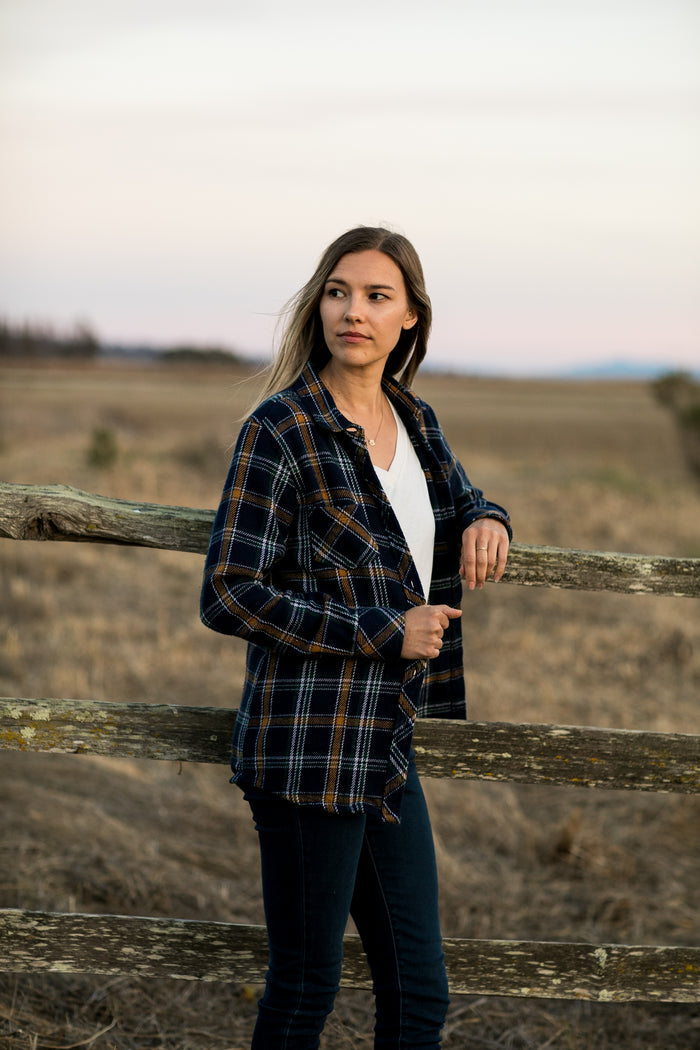 A woman wears the Pladra Women's Fireside Flannel in Feather Blue, standing by a fence at sunset.