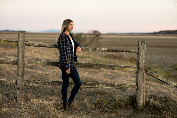 A woman wears the Pladra Women's Fireside Flannel in Feather Blue by a rustic fence in a sunlit open field.