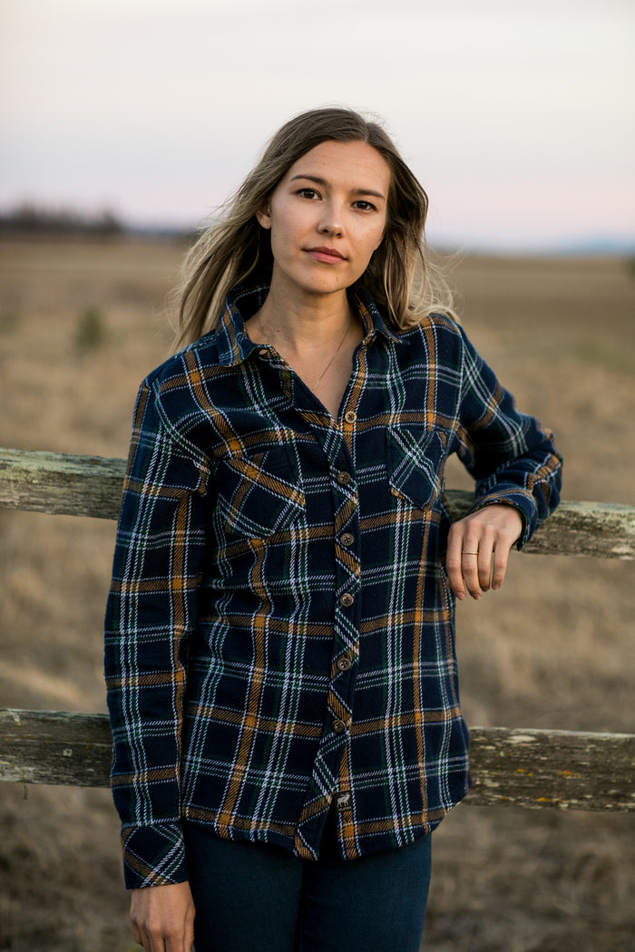 A woman wears the Pladra Women's Fireside Flannel in Feather Blue by a wooden fence in a grassy field at sunset.