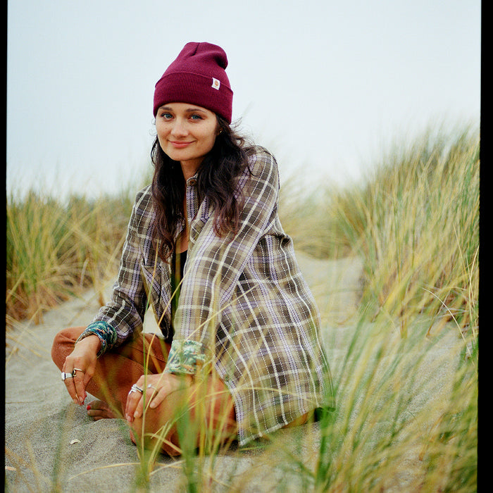 A woman in a maroon beanie, rings, and the Women's Fireside Flannel in Grove Green sits cross-legged on sandy ground amid tall grass, smiling softly at the camera under an overcast sky.