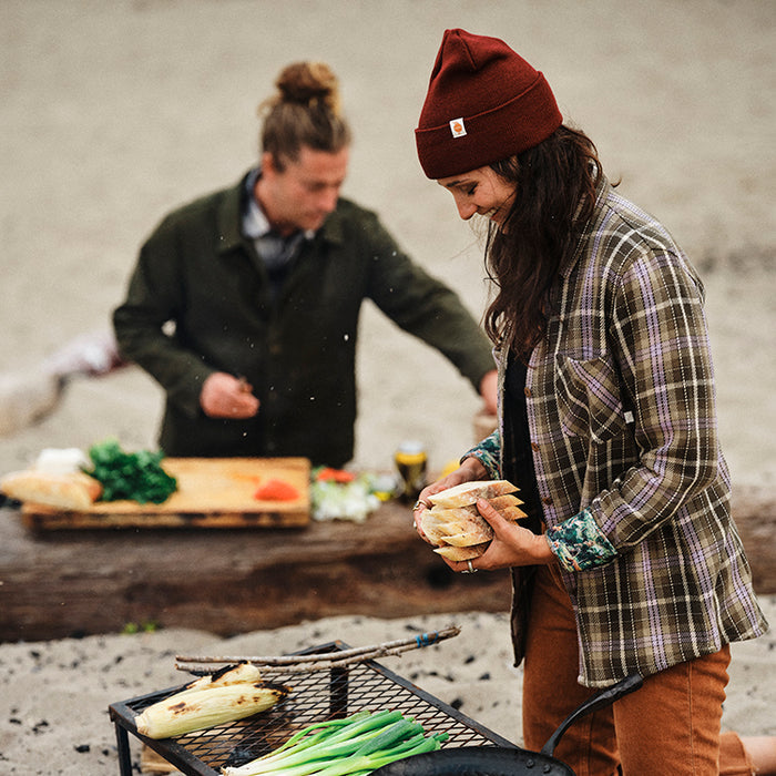 A woman in a Pladra Women's Fireside Flannel - Grove Green grills outdoors beside a man chopping vegetables.