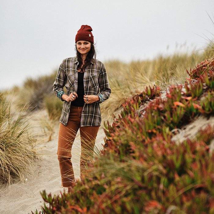 Woman in Pladra's Women's Fireside Flannel–Grove Green stands on a sandy beach with dune grass and coastal plants.