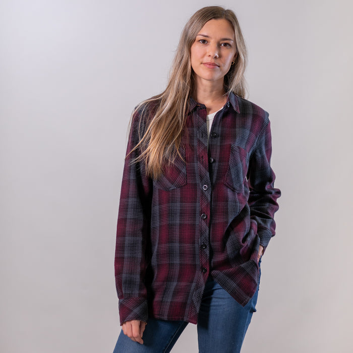 A woman with long brown hair wears the Women's Fireside Flannel - Hickory Red and blue jeans, standing against a plain light background with one hand in her pocket and a neutral expression.