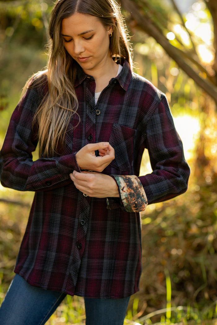Outdoors in the sunlight, a woman adjusts the sleeve of her Women's Fireside Flannel - Hickory Red, featuring heavyweight burgundy and black plaid with rolled cuffs. Blurred trees and greenery fill the background.