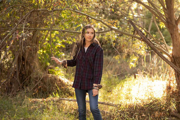 A young woman stands outdoors in a sunlit, wooded area, gently holding a tree branch and looking thoughtfully to the side while wearing the Women's Fireside Flannel- Hickory Red and jeans.