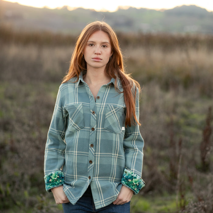 A young woman with long red hair stands in a field, wearing the Women's Fireside Flannel - Island Blue over blue jeans, looking directly at the camera with a neutral expression.