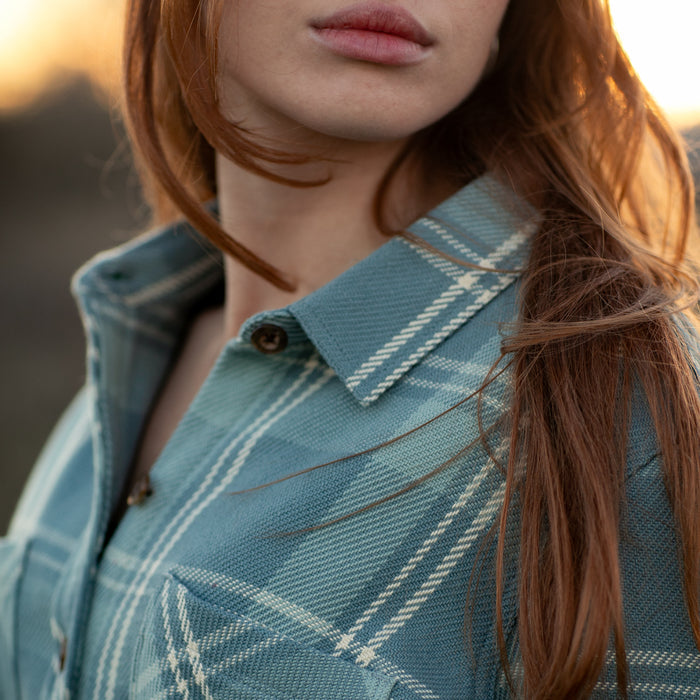 A close-up of a woman with long red hair wearing the Women's Fireside Flannel in Island Blue, made from organic cotton flannel. She stands outdoors in warm sunlight, her lips and chin partially visible.
