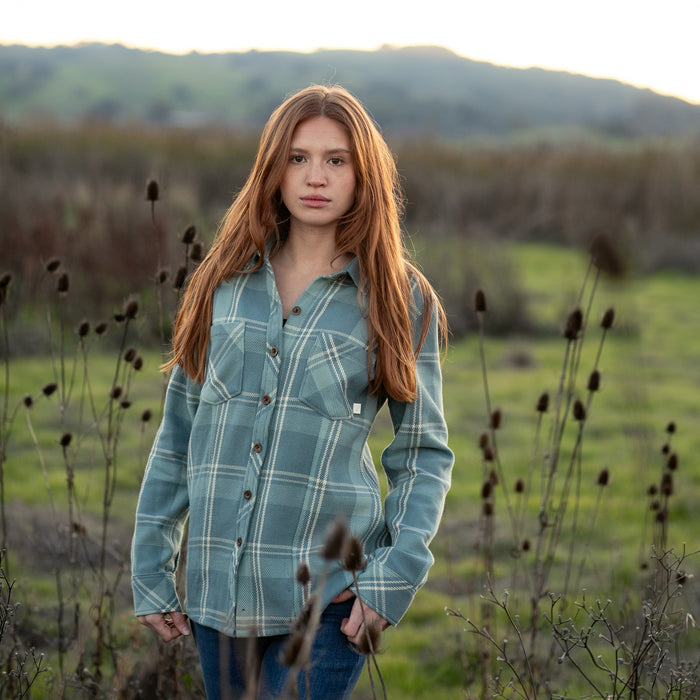 A young woman with long red hair, wearing the Women's Fireside Flannel in Island Blue and jeans, stands in a field of dry plants with green hills behind her.