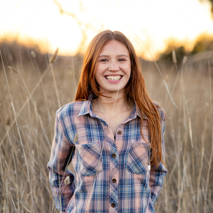 Smiling young woman in a Pladra Women's Fireside Flannel - Lily Pink stands in a sunlit grassy field.