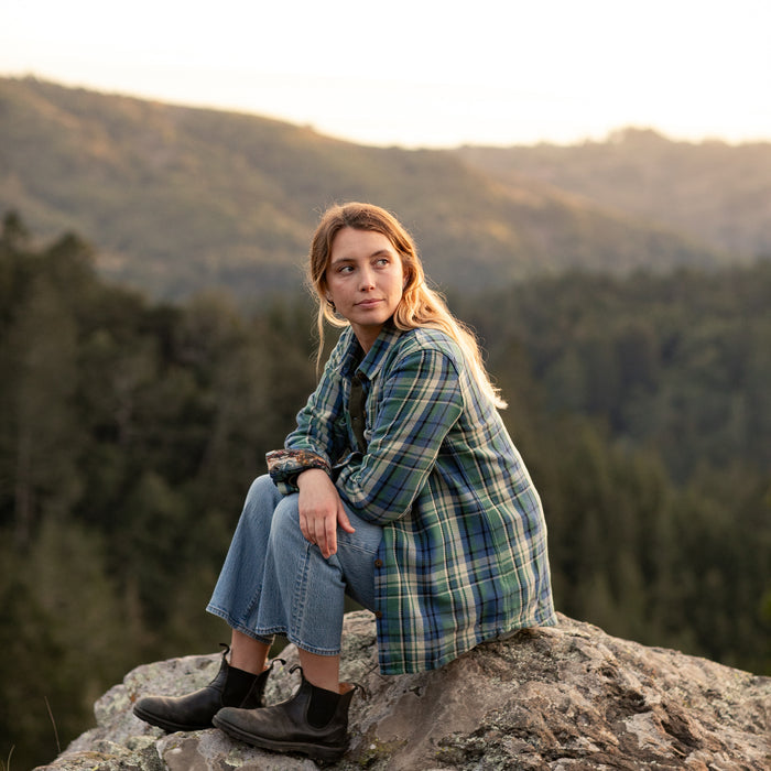 Wearing the Women's Fireside Flannel in Primrose Green, a woman with long hair and jeans sits on a rock before forested hills at sunset, gazing thoughtfully into the distance.