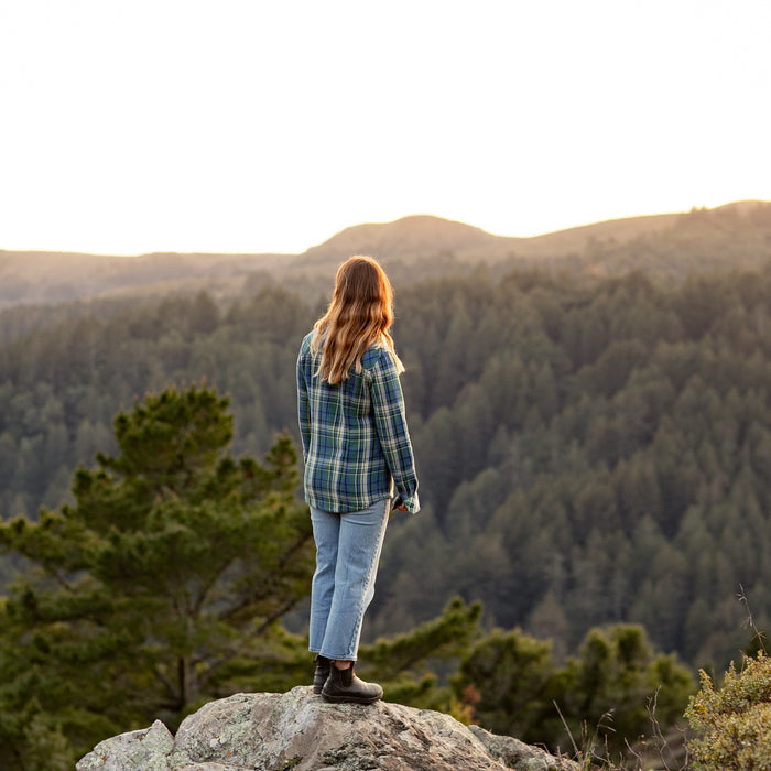 Wearing the Women's Fireside Flannel in Primrose Green and jeans, a woman with long hair stands on a rocky outcrop, gazing over forested hills at sunset.