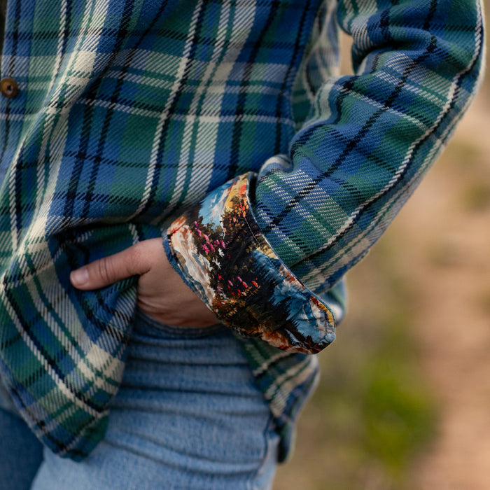 A woman wears the Women's Fireside Flannel in Primrose Green, its blue and green plaid with rolled-up sleeves showing a colorful lining, paired with blue jeans, as she stands with one hand in her pocket.
